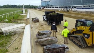 Landscaping at a race track is underway with large boulders, a dump truck, and a skid steer loader.