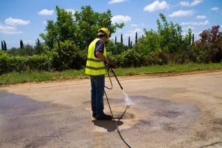 A worker in safety gear uses a power washer to clean a pavement surface.