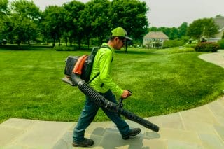 A landscaper, wearing a bright green shirt, uses a backpack leaf blower near a green lawn.